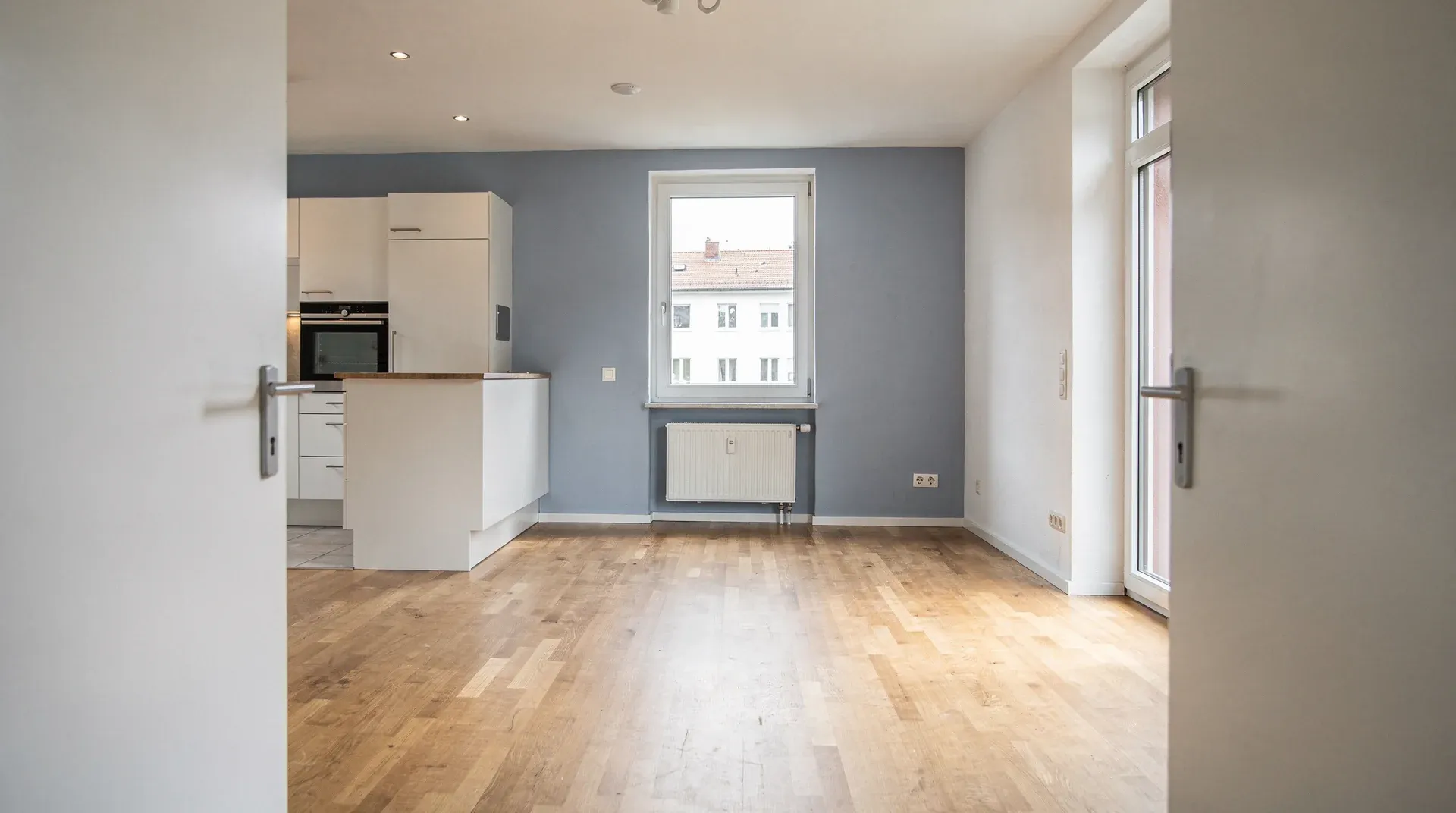 Leere Wohnung mit Einbauküche, Eichenparkett und blaugrauer Wand, Blick durch die geöffnete Zimmertür